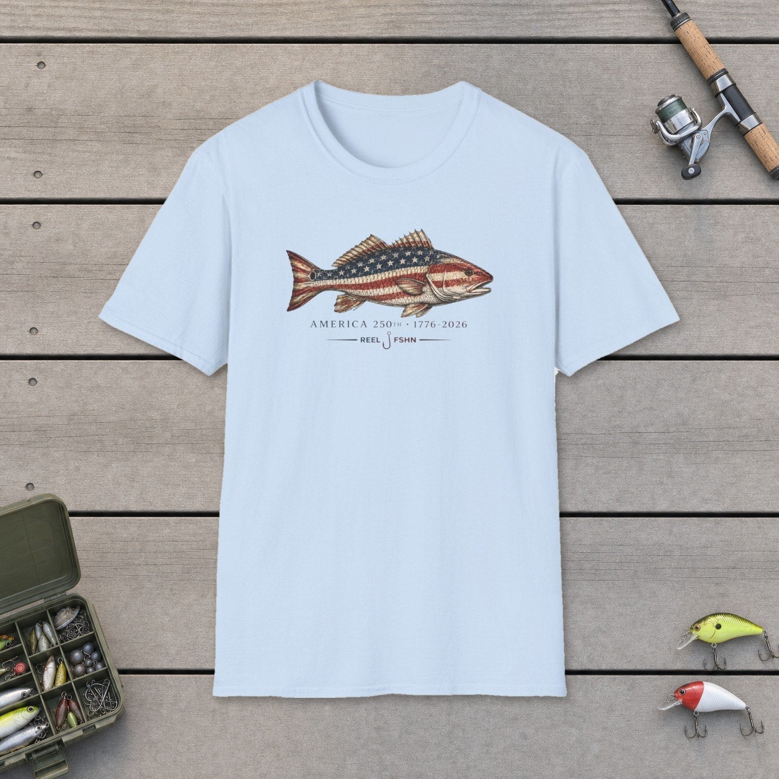 America's 250th T-Shirt flat-laid on wooden dock boards beside a fishing rod and tackle box